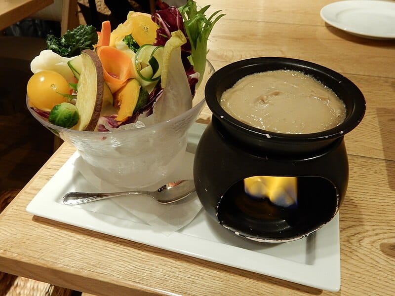 Rechaud com fondue e uma tijela de vegetais sobre um prato branco com uma colher que está sobre a mesa da madeira