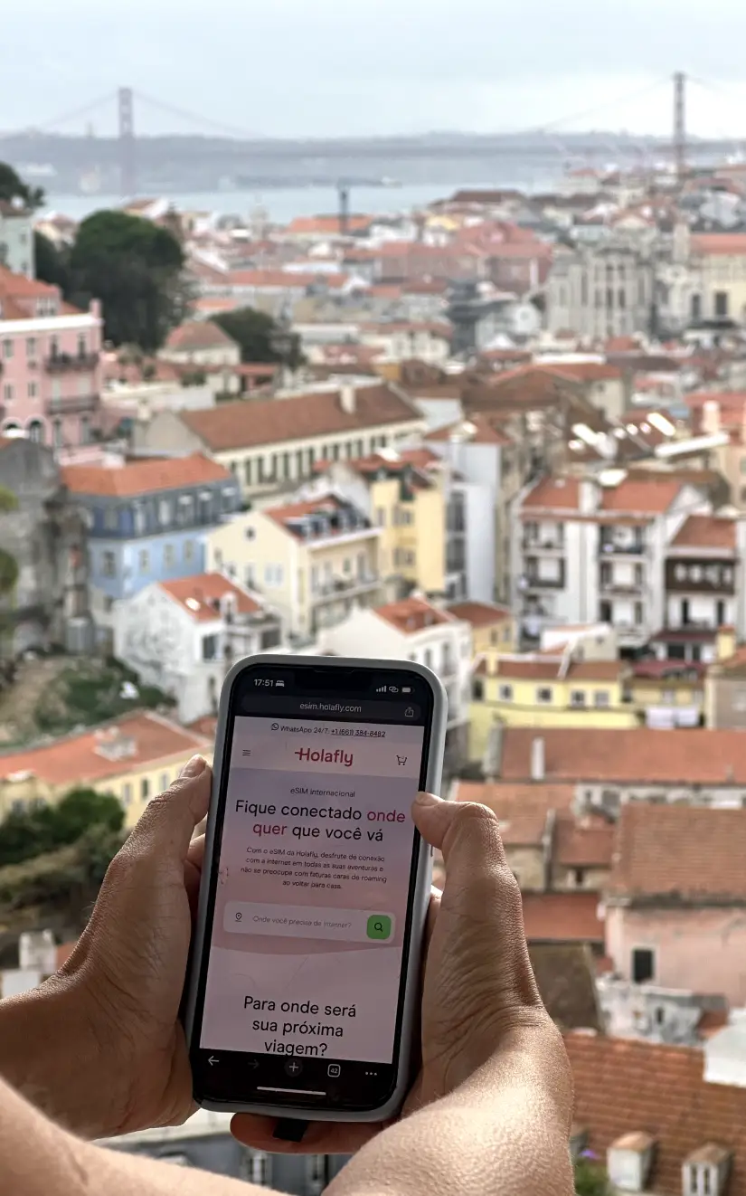 Uma pessoa segurando um telefone captura os telhados de Lisboa e a icônica ponte ao fundo, combinando o charme e a conectividade da Holafly Portugal.