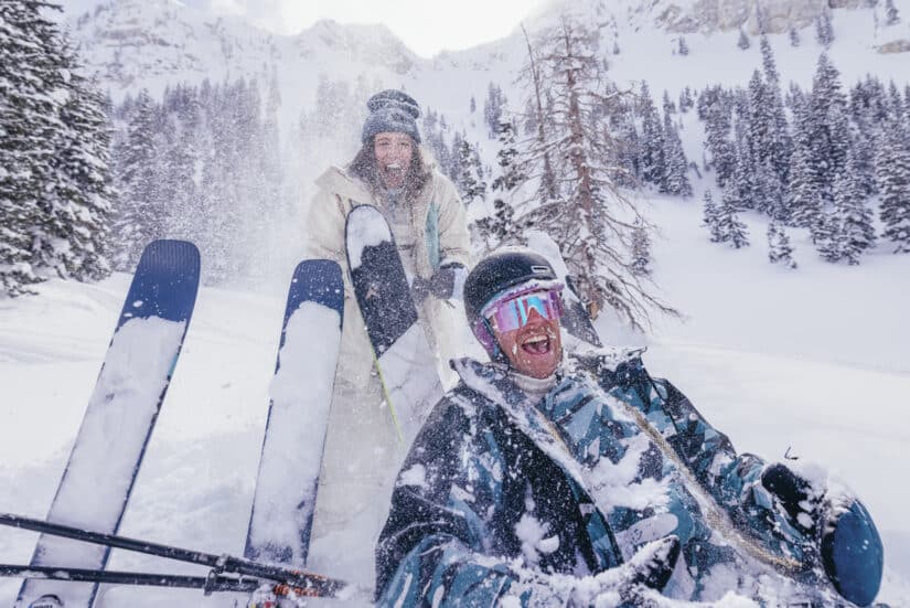 Duas pessoas aproveitam um dia de neve esquiando com roupas de ski, uma delas sentada rindo em meio à neve e aos pinheiros.