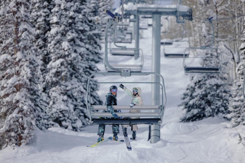 Dois esquiadores em roupa de esqui acenam de um teleférico coberto de neve, cercados por árvores cobertas de neve em um dia de inverno.