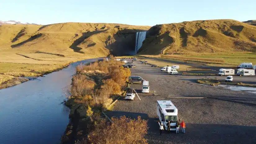 Vista aérea de campistas perto de um rio, com a cachoeira Skogafoss ao fundo e Rv's em colinas abertas e gramadas.