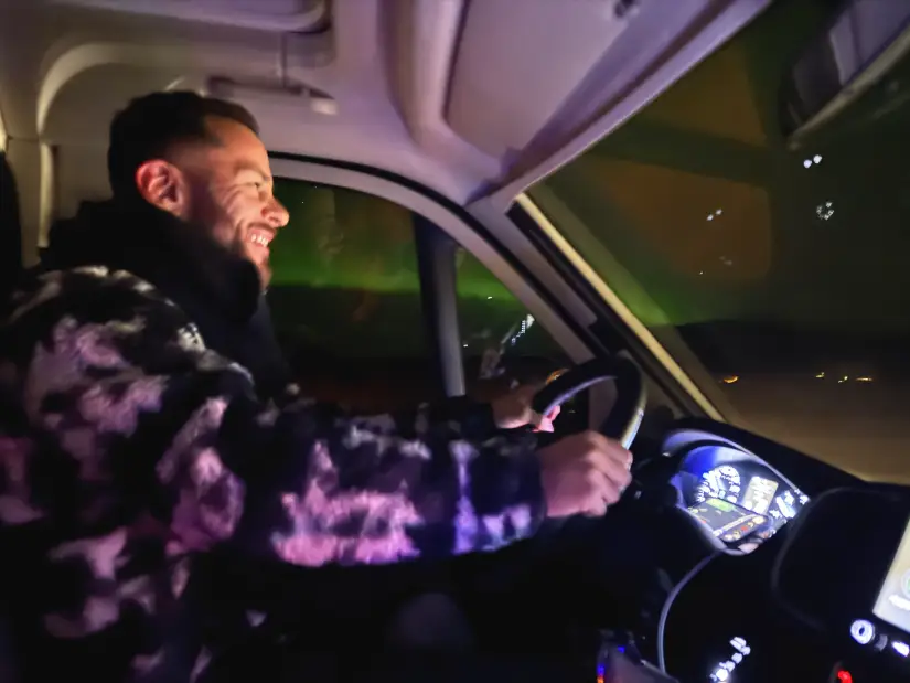 Homem sorrindo enquanto dirige um veículo à noite, com luzes da aurora boreal do lado de fora da janela do veículo.