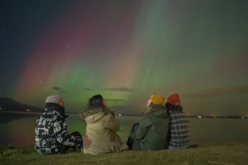 Quatro pessoas sentadas em gramado em frente a um lago, curtindo as luzes da aurora boreal.