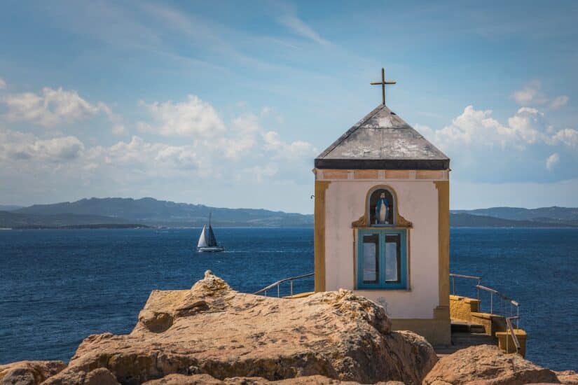 Rochas com uma torre de igreja em destaque, com a vista para o mar com um barco a vela