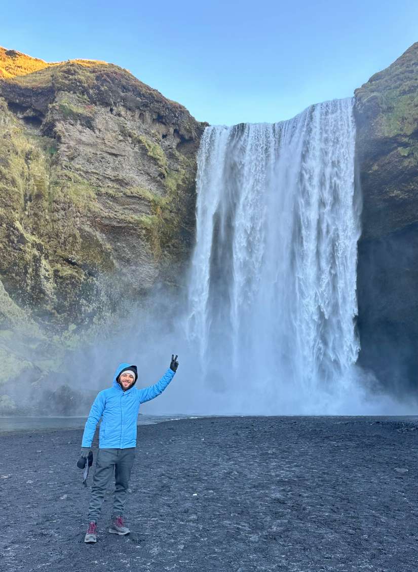 Pessoa de jaqueta azul impermeável da Columbia, item essencial para levar na mala para a Islândia, posa em frente a uma grande cachoeira em um dia claro.