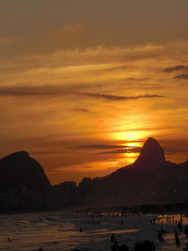 O pôr do sol sobre uma praia animada com montanhas recortadas contra um céu alaranjado é semelhante aos deslumbrantes passeios no Rio de Janeiro, onde paisagens vibrantes criam momentos de tirar o fôlego.