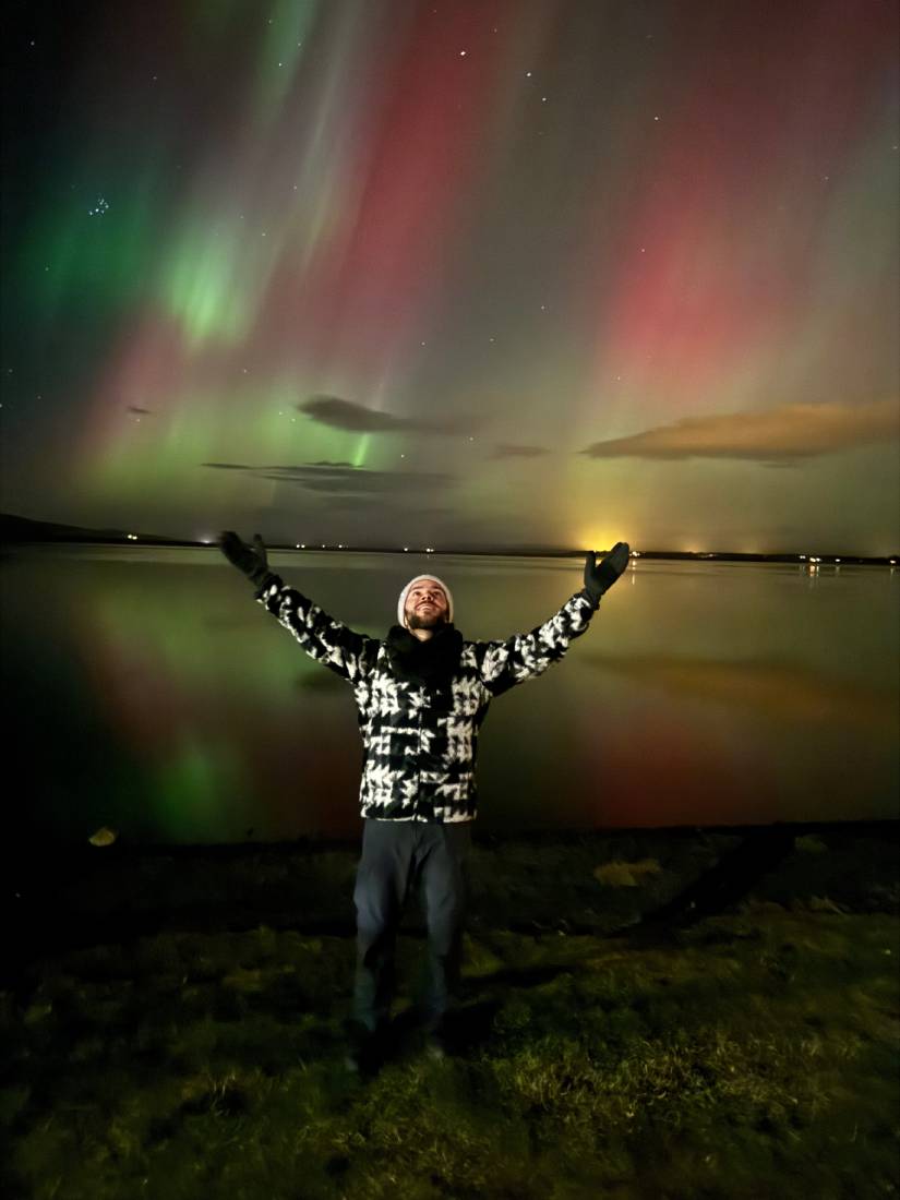 Pessoa alegremente parada sob a vibrante aurora boreal na Islândia, perto de um lago tranquilo à noite.