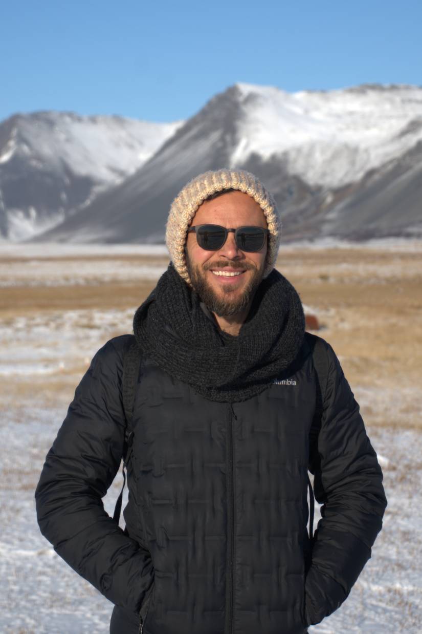 Uma pessoa com roupas de inverno sorri ao ar livre com montanhas cobertas de neve na Islândia.