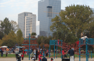 Crianças brincando em brinquedos de playground em um parque, com prédios altos ao fundo.