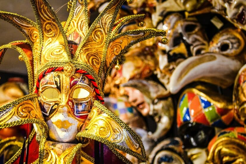 Uma máscara de carnaval vibrante e ornamentada com detalhes dourados e pontas semelhantes às de um bobo da corte, entre outras máscaras coloridas.