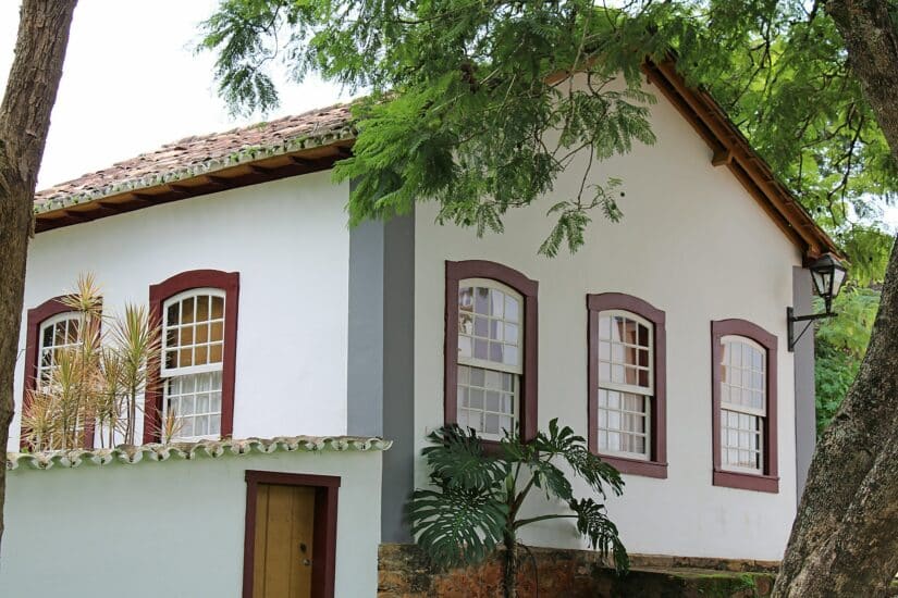 Uma casa em estilo colonial, branca e vermelha, com várias janelas, está aninhada entre as árvores, evocando o Carnaval no interior de Minas.