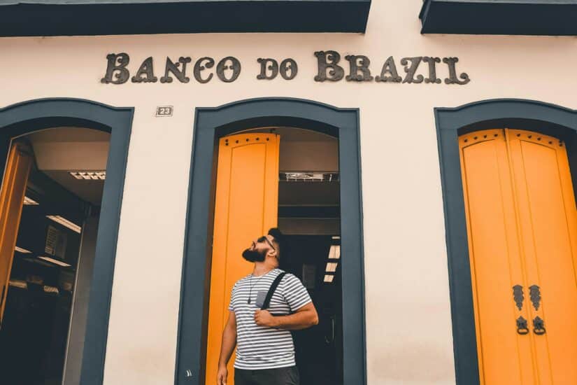 Um homem de camisa listrada está em frente ao Banco do Brasil, com portas com detalhes em laranja.