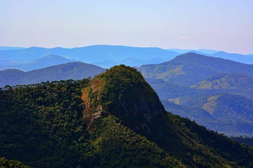 Pico verdejante de montanha semelhante ao Carnaval no interior de Minas, cercado por distantes colinas azuis sob um céu limpo.