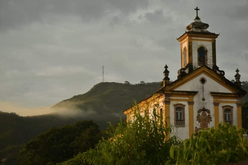 Uma igreja histórica com detalhes ornamentados se destaca em um cenário de colinas verdejantes e um céu nublado.