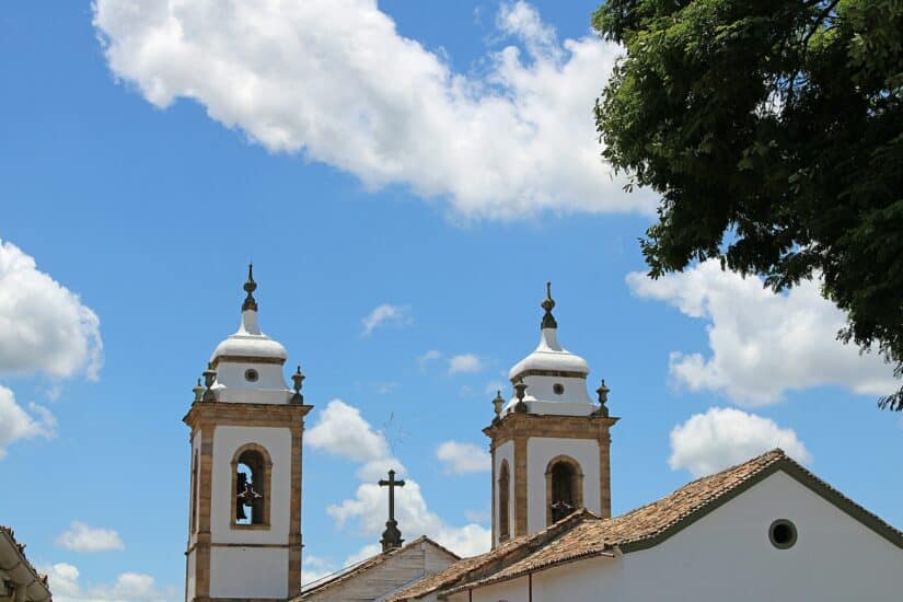 Igreja histórica com duas torres sineiras sob um céu de Carnaval no interior de Minas e uma árvore à direita.