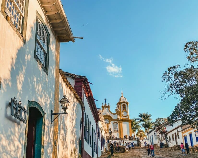 Cena de rua colonial em Tiradentes, Brasil, com edifícios vibrantes, uma igreja distante e o espírito do Carnaval no interior de Minas.